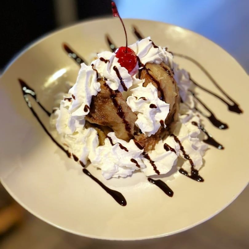 Fried ice cream dessert with whipped cream, chocolate drizzle, and a cherry on a white plate.