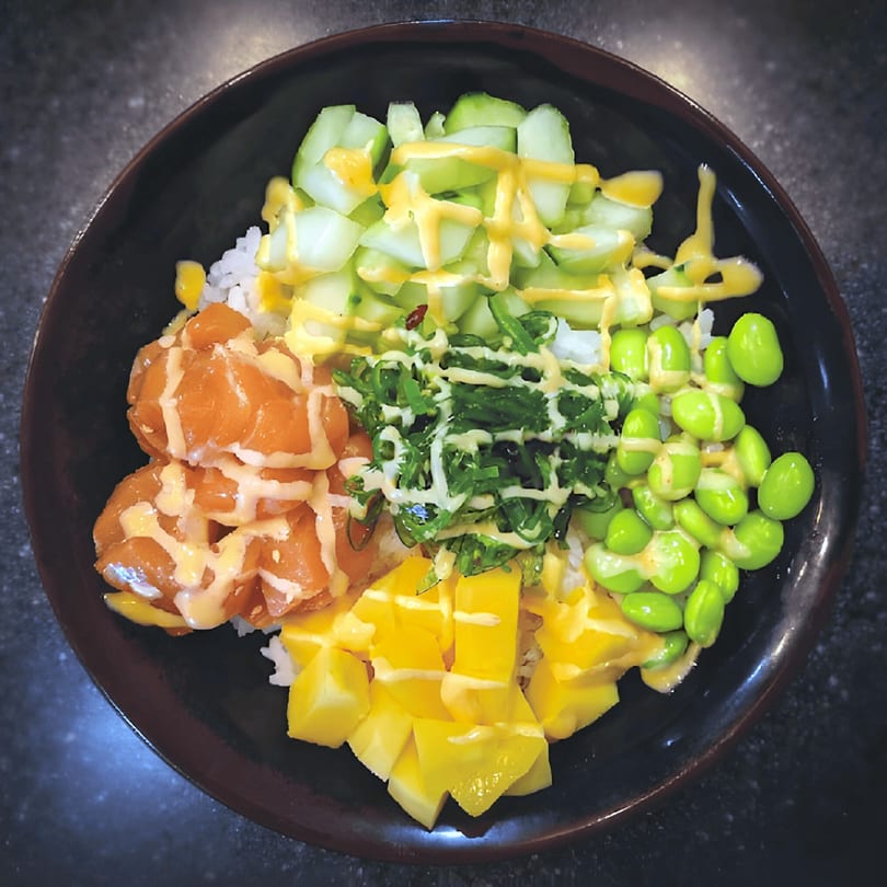 Top-down view of a colorful Japanese poke bowl with salmon, mango, edamame, and cucumber.